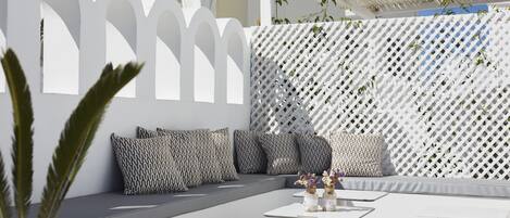 Un espacio al aire libre moderno con mobiliario blanco y cojines estampados, rodeado por una cerca de celosía blanca y un edificio blanco al fondo.