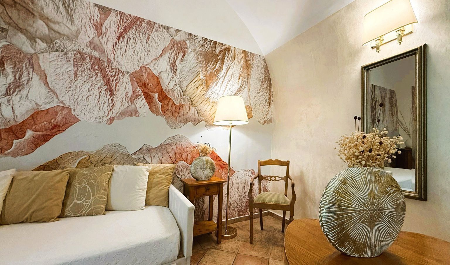 A bedroom with a bed, a chair, a lamp, a vase with flowers, and a wall with a mountain mural.