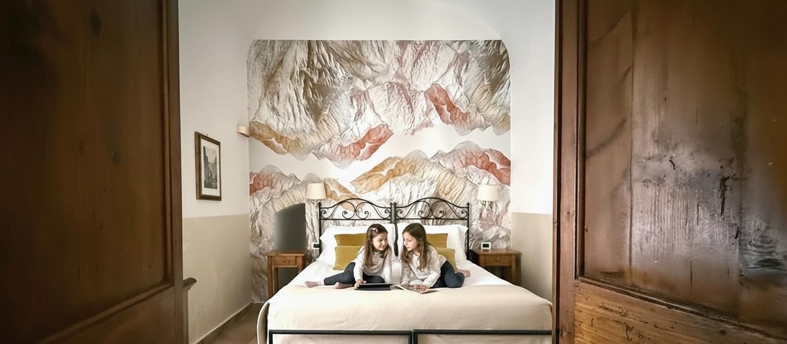 Un dormitorio con una cama, dos niños sentados encima, una mesita de noche y un mural en la pared.