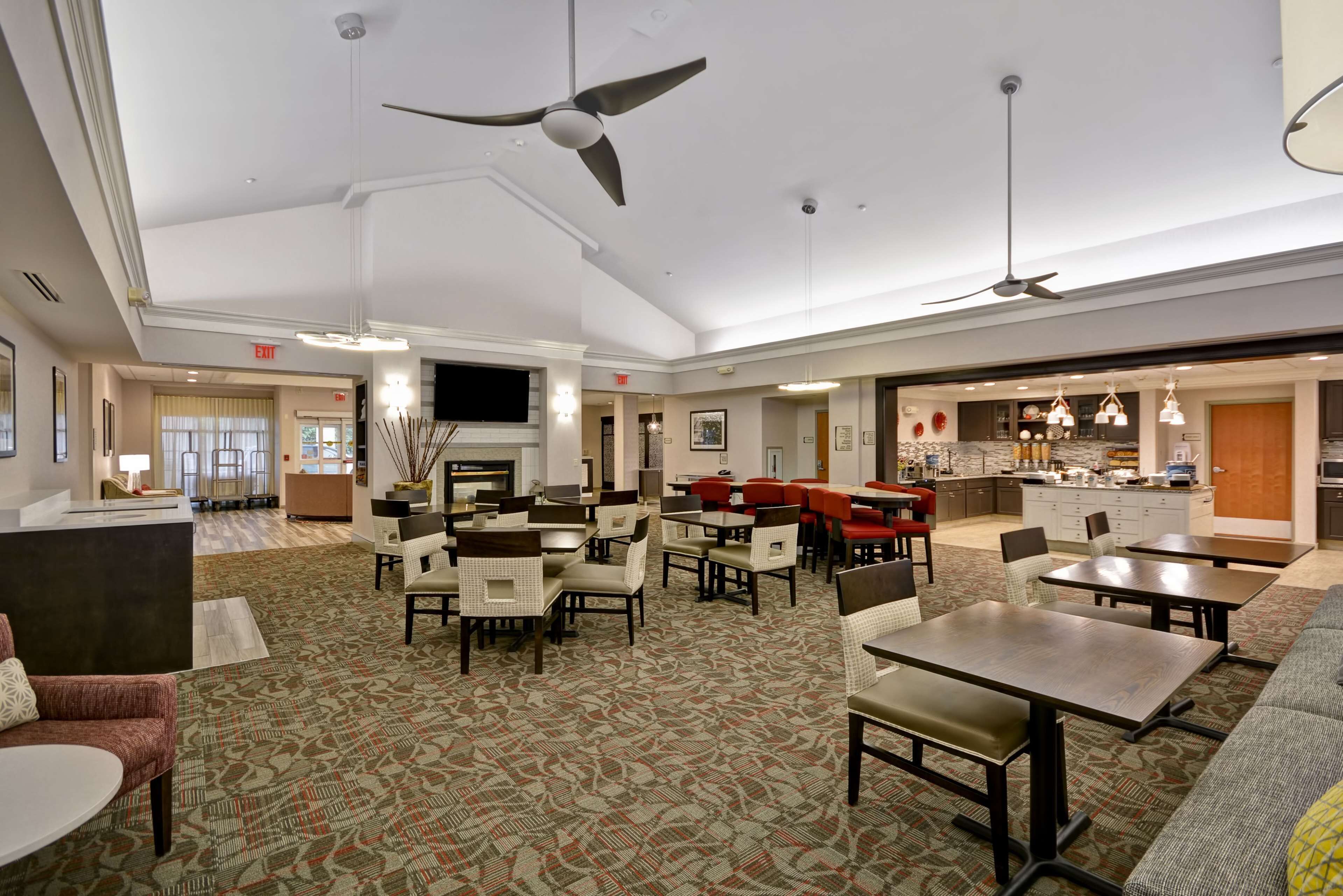A spacious hotel lobby with multiple seating areas, a kitchen in the background, and ceiling fans.