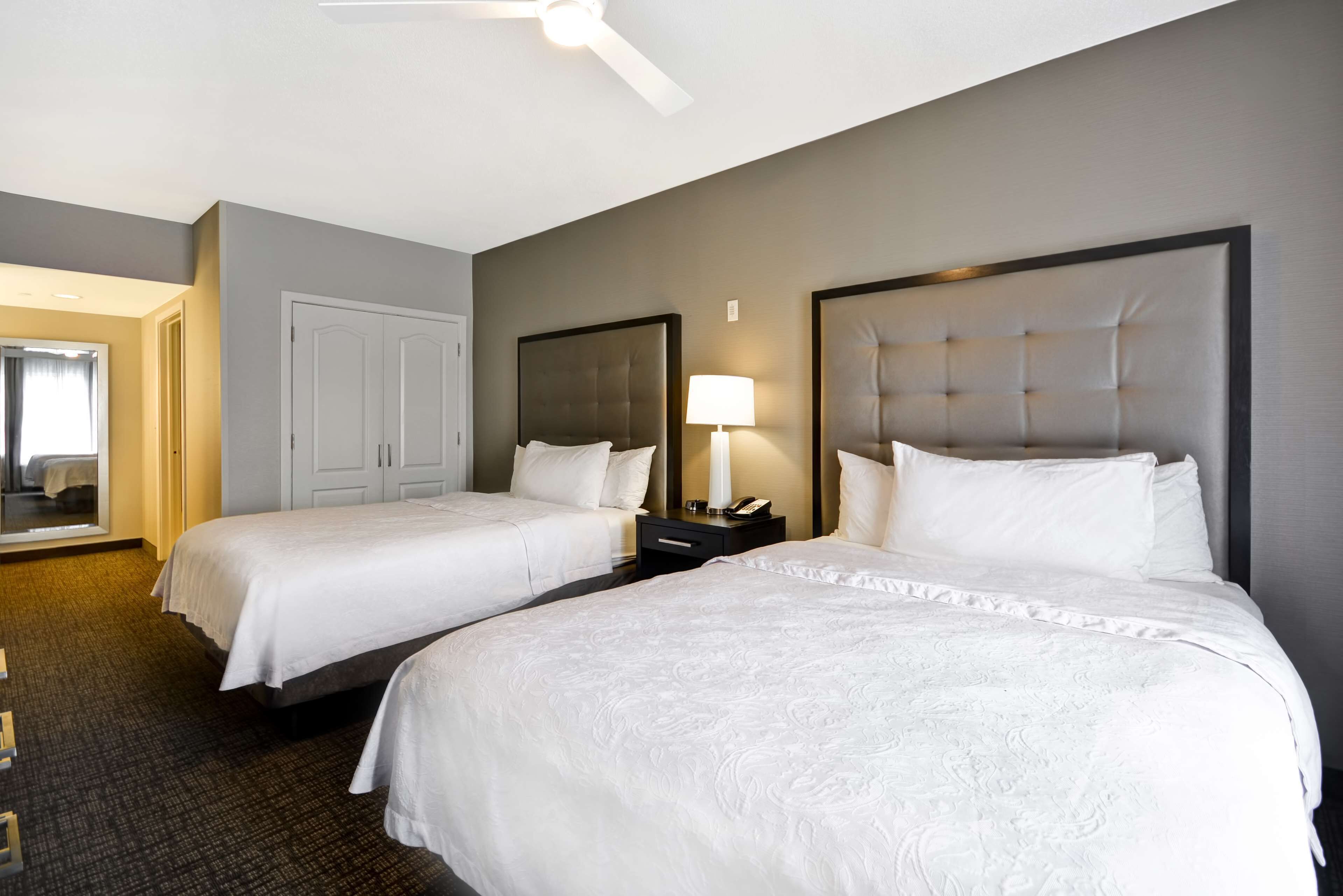 A hotel room with two beds, a ceiling fan, a nightstand with a lamp, and a wardrobe.