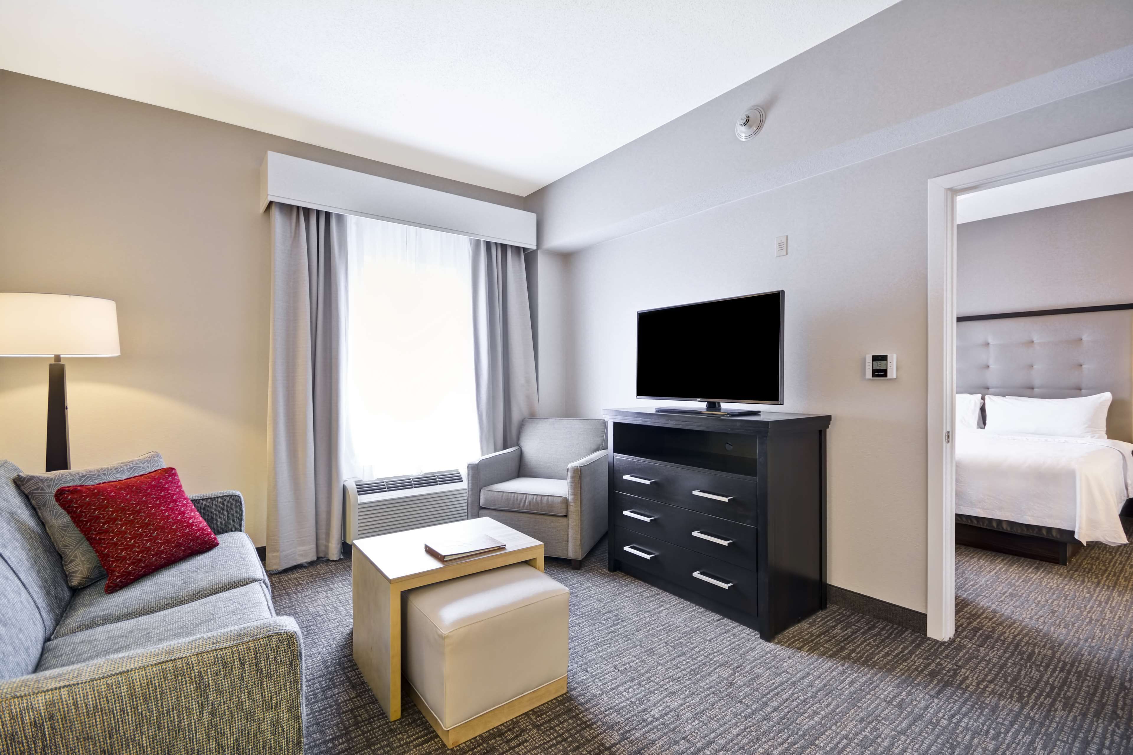 Ett hotellrum med en grå soffa, en röd kudde, en vit pall, en platt-TV på ett svart stativ, en grå fåtölj, en vit säng och en grå matta.