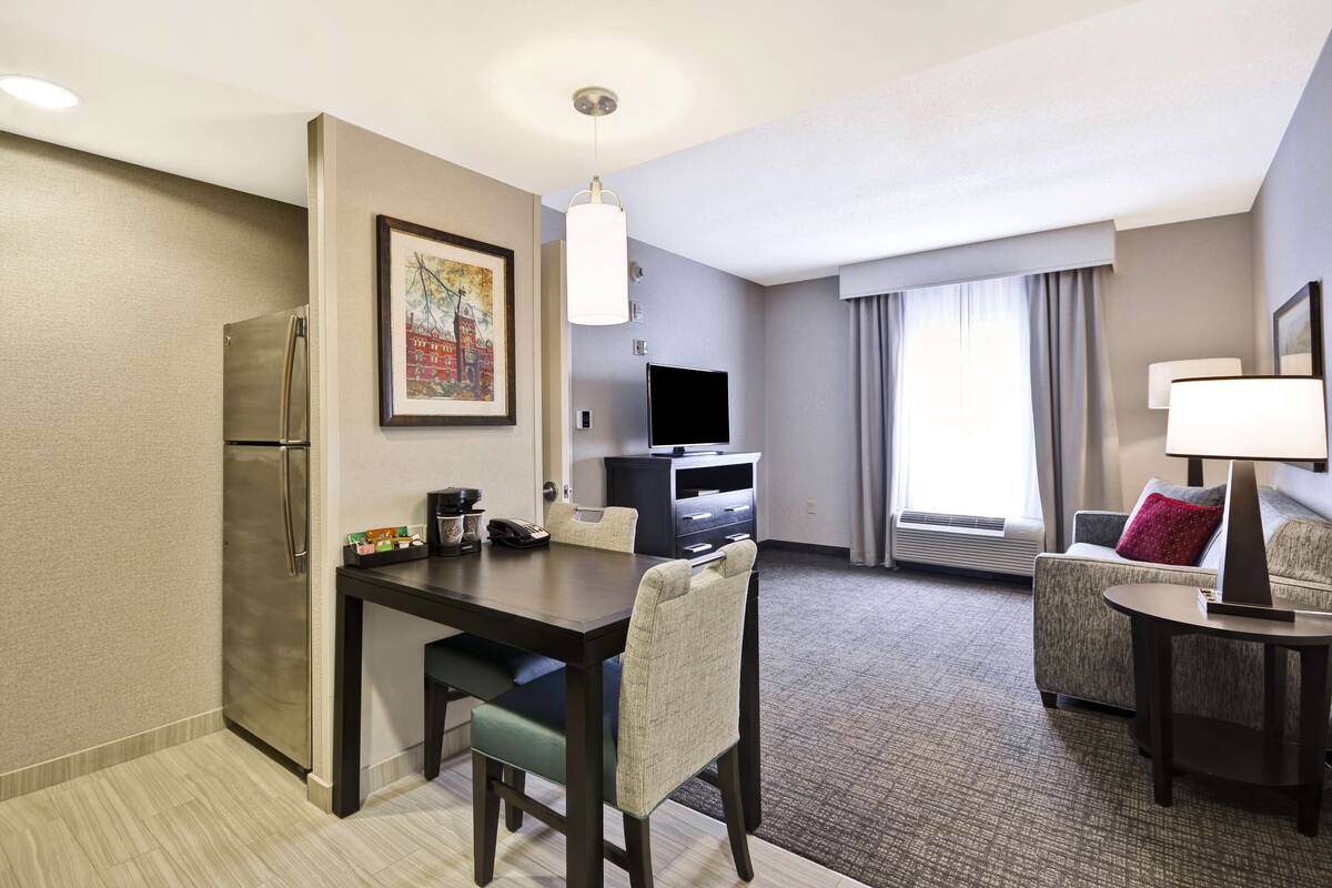 Quarto de hotel com cozinha compacta, área de jantar, espaço de estar com sofá e poltrona, televisão de tela plana e uma janela grande com cortinas.
