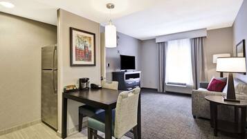 Habitación de hotel con kitchenette, comedor, zona de estar con sofá y sillón, televisor de pantalla plana y un amplio ventanal con cortinas.