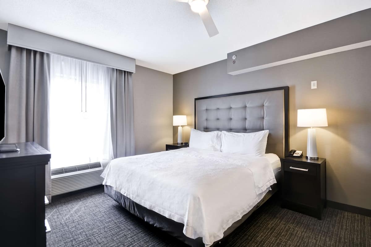 Quarto de hotel com uma cama grande, duas mesas de cabeceira com luminárias, um ventilador de teto e uma janela ampla com cortinas.