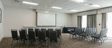 Un salón de conferencias con filas de sillas, una pantalla de proyector y una mesa.