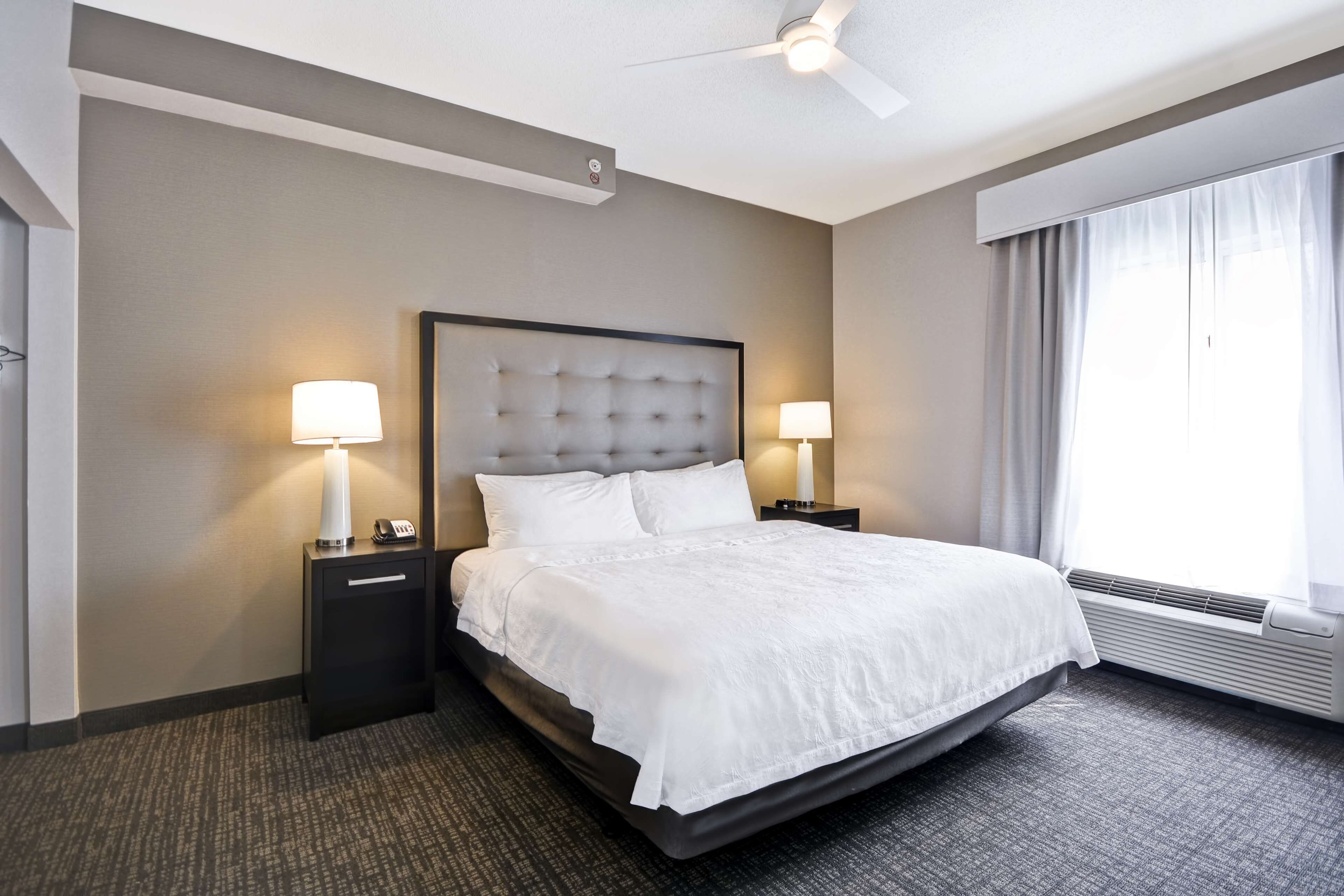 A hotel room with a large bed, two bedside tables with lamps, a ceiling fan, and a window with curtains.