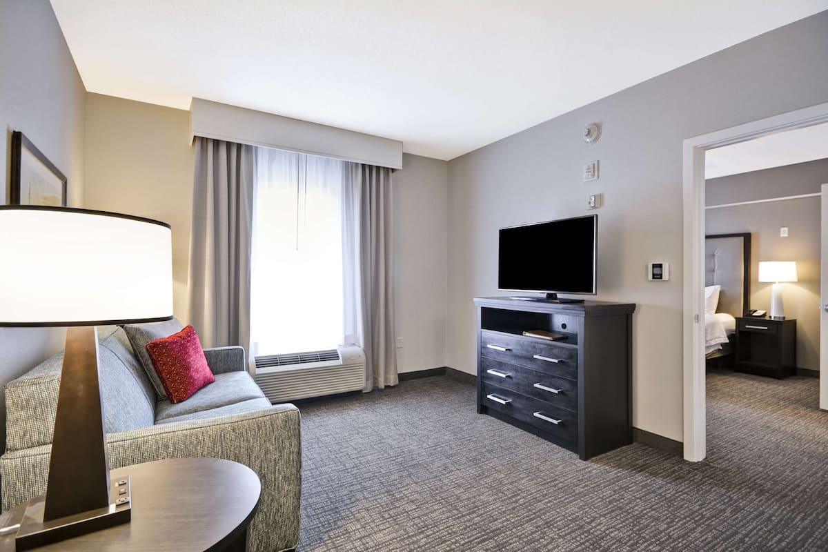 Quarto de hotel com um sofá cinzento, uma televisão de tela plana sobre uma cômoda escura, um abajur e uma janela com cortinas.