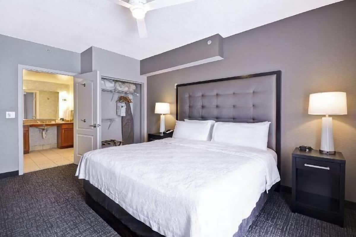 Quarto de hotel com uma cama grande, duas mesas de cabeceira com luminárias, uma porta que dá para um banheiro e uma pia de banheiro visível.