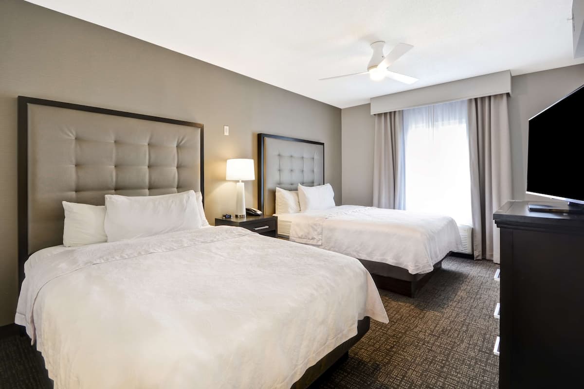 Quarto de hotel com duas camas, ventilador de teto, televisão e janela com cortinas.