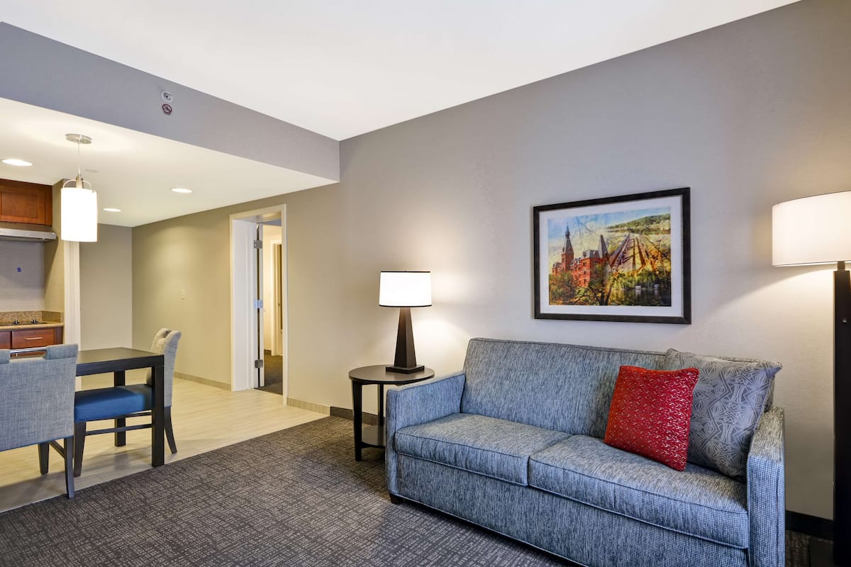 Quarto de hotel com área de jantar, um sofá com almofada vermelha, uma luminária e um quadro na parede.