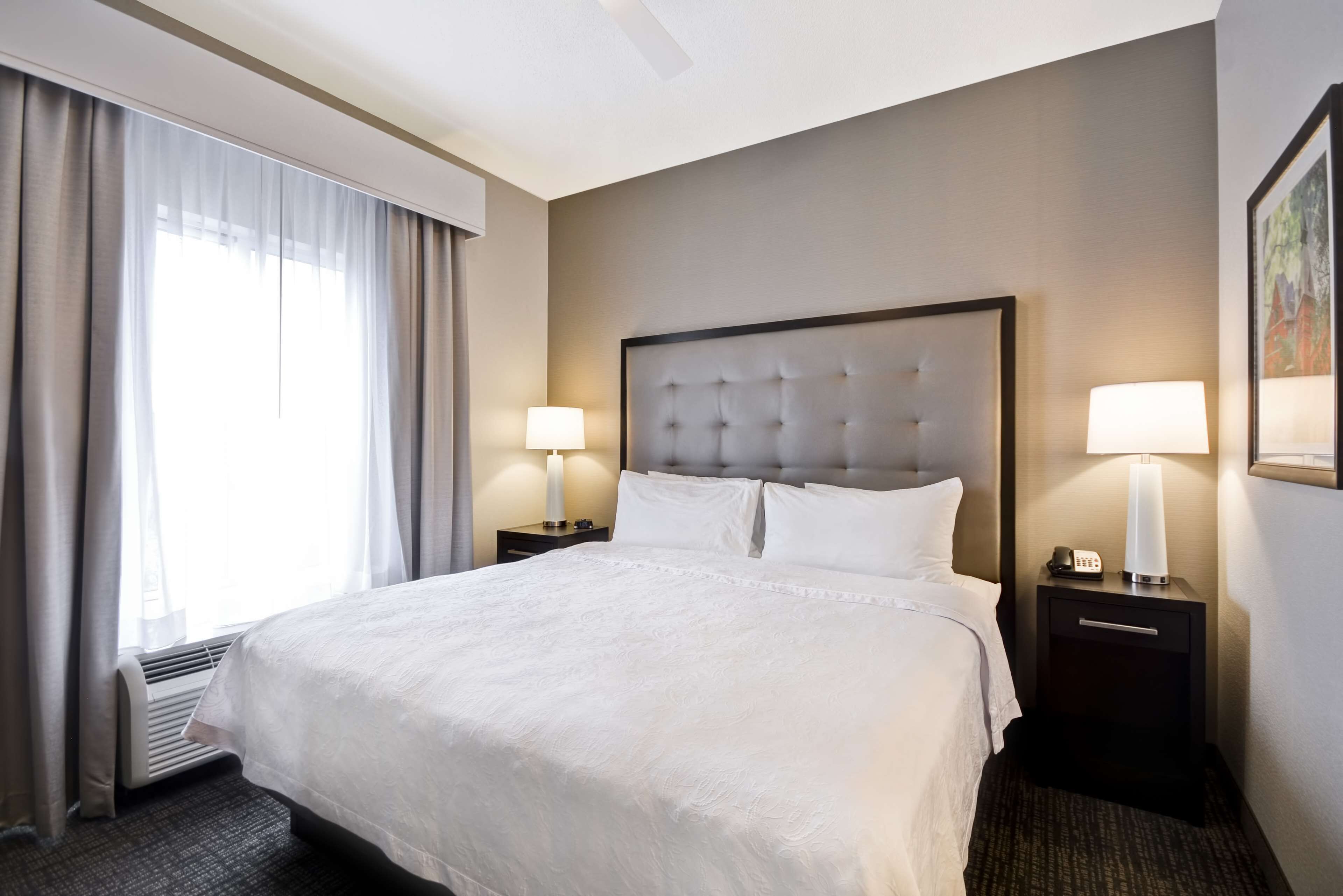 Quarto de hotel com uma cama grande, duas mesas de cabeceira com luminárias, uma janela com cortinas e uma obra de arte na parede.