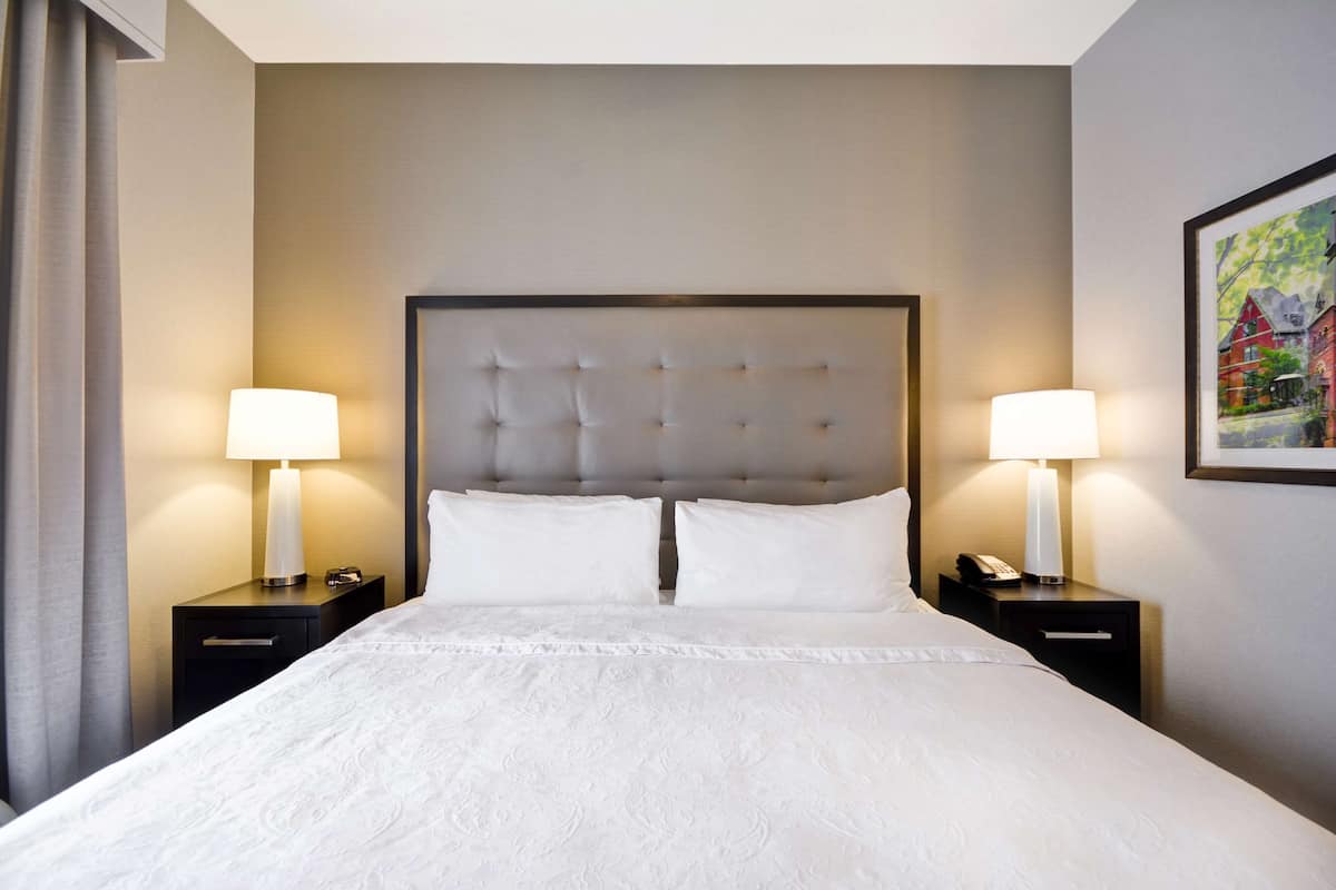 Quarto de hotel com uma cama grande, duas mesas de cabeceira com abajures, cabeceira e um quadro na parede.