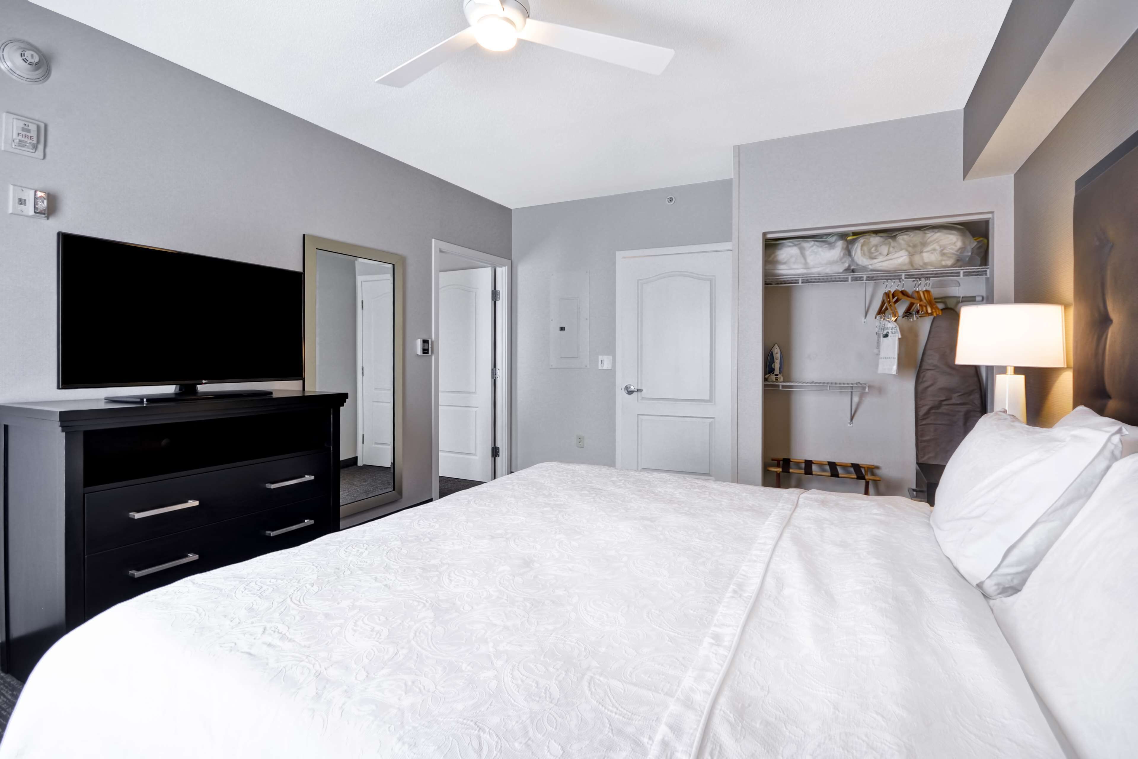 A hotel room with a bed, a TV on a dresser, a closet with hanging clothes, and a ceiling fan.