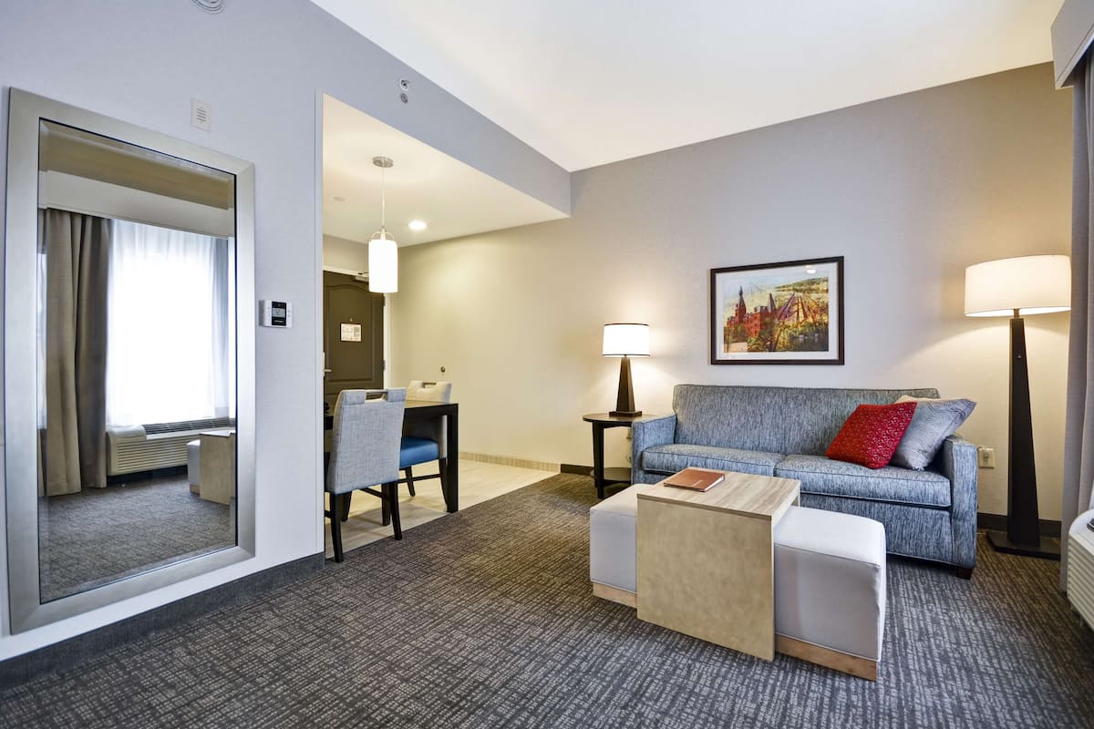 Quarto de hotel com carpete cinza, sofá azul com almofadas vermelhas e brancas, mesa de centro de madeira, mesa lateral com abajur e um quadro emoldurado na parede.