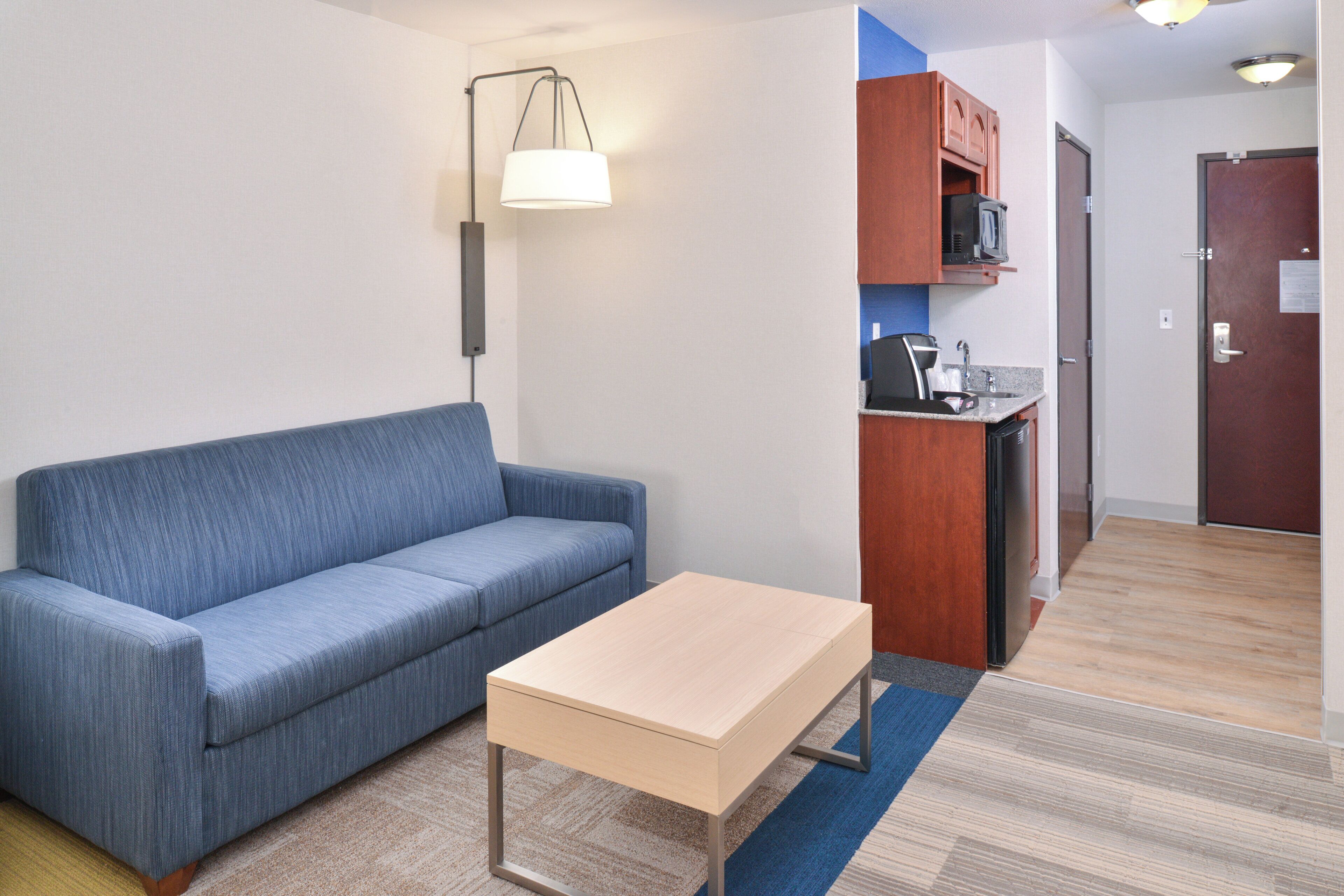 Un espacio habitable compacto con sofá, una mesita, kitchenette y una puerta que da a otra habitación.