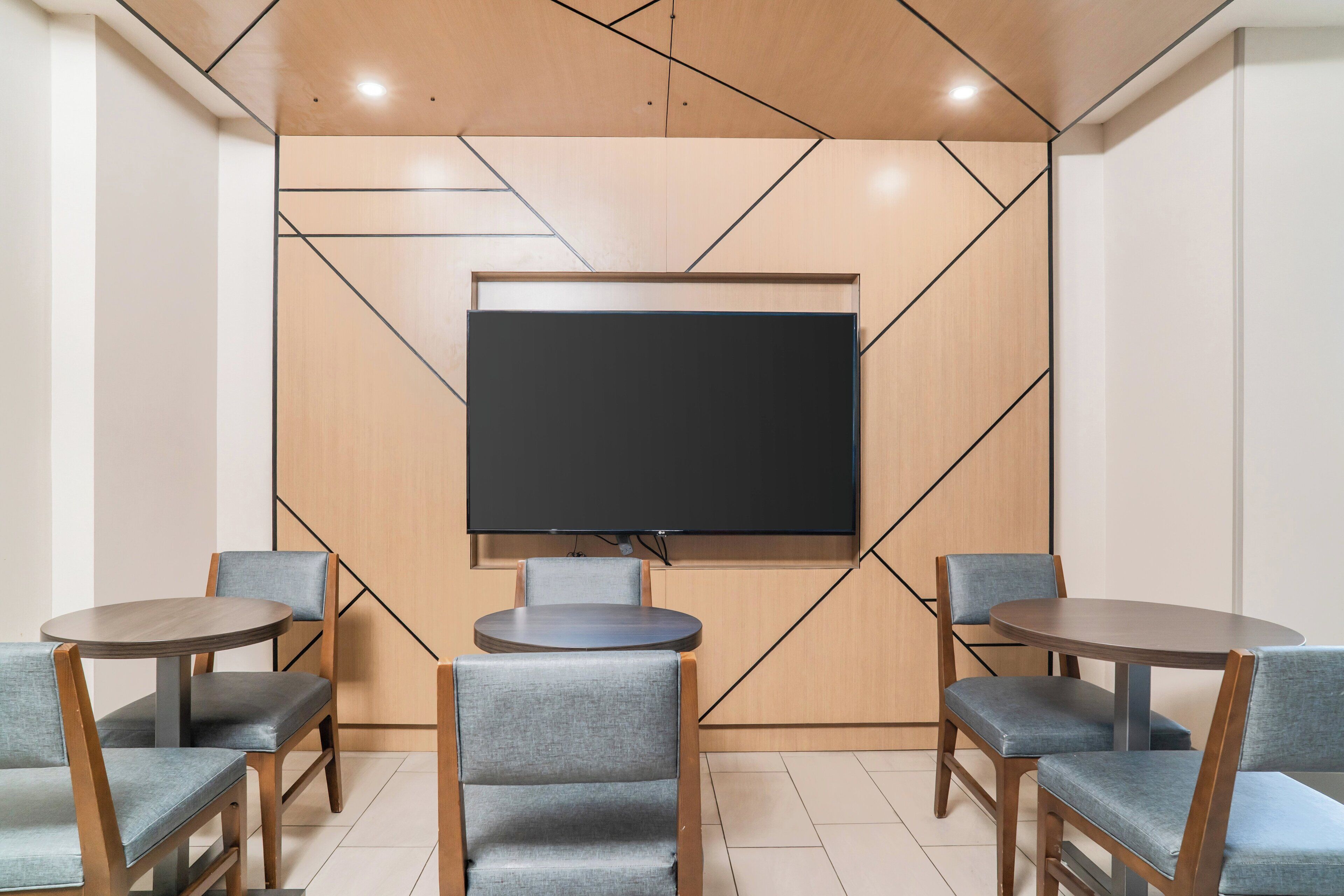 A conference room with a large flat-screen TV, wooden panel wall design, and four grey upholstered chairs around two round tables.