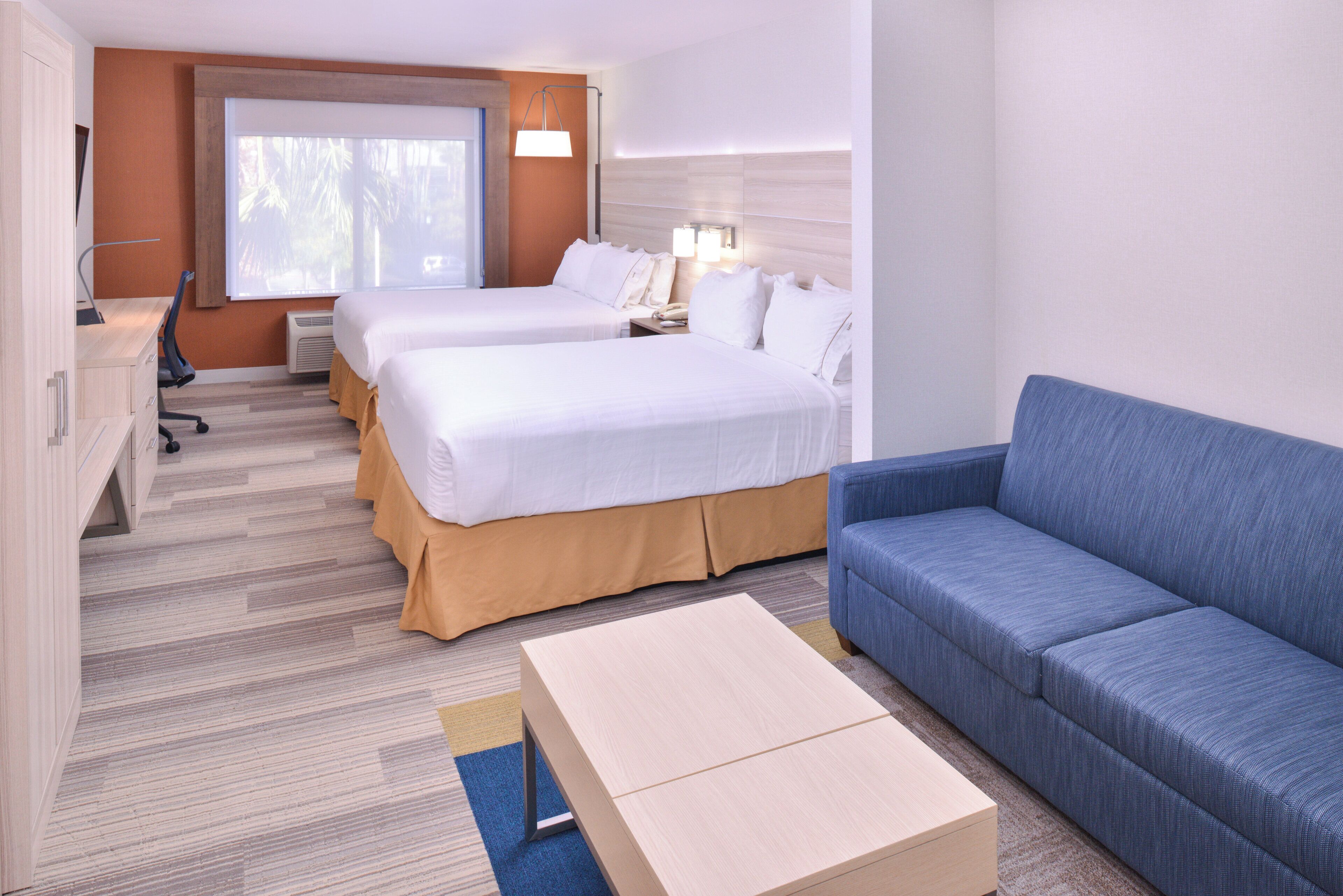 Ein Hotelzimmer mit zwei Betten, einem Schreibtisch, einem Sessel und einer Couch, sowie einem Fenster mit Jalousien.