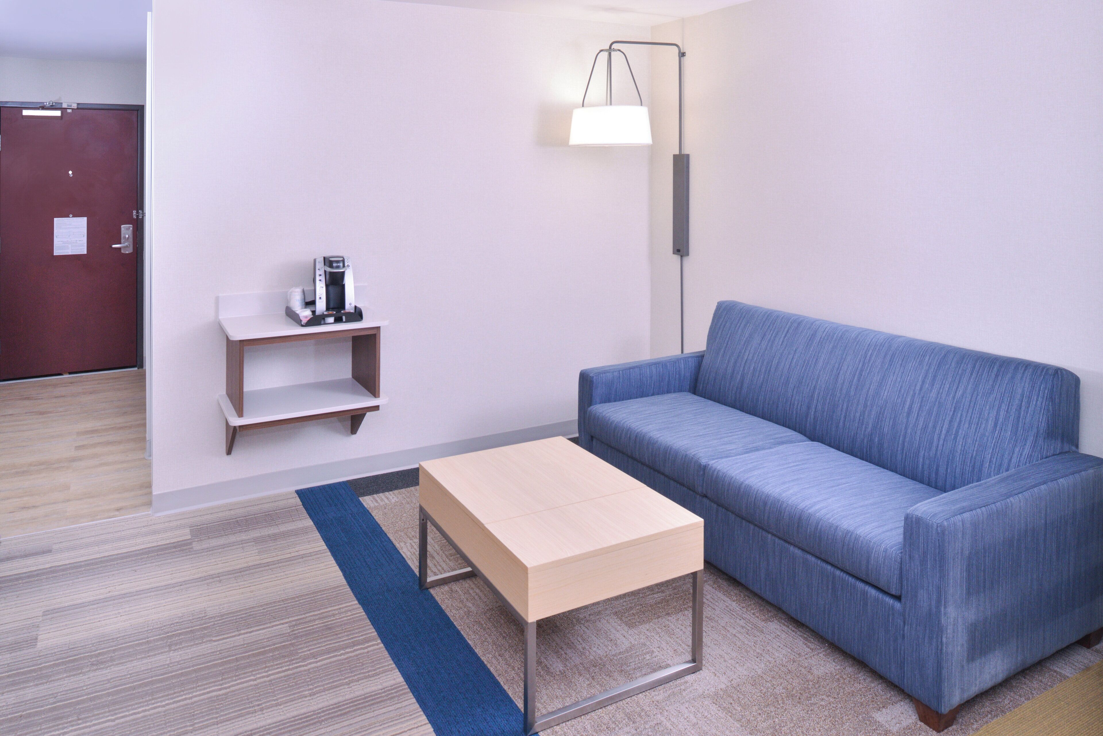 A room with a blue sofa, a small wooden table, a lamp, and a coffee machine on a shelf.