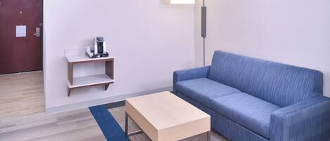 A room with a blue sofa, a small wooden table, a lamp, and a coffee machine on a shelf.