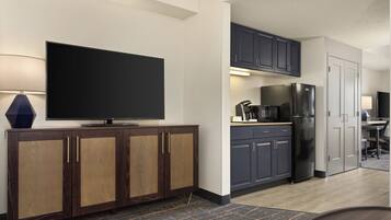 A modern kitchen with dark cabinets, a large flat-screen TV, a refrigerator, and a coffee maker.