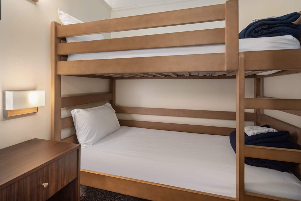 Una cama litera con estructura de madera, ropa de cama blanca y una mesita de noche con cajón.