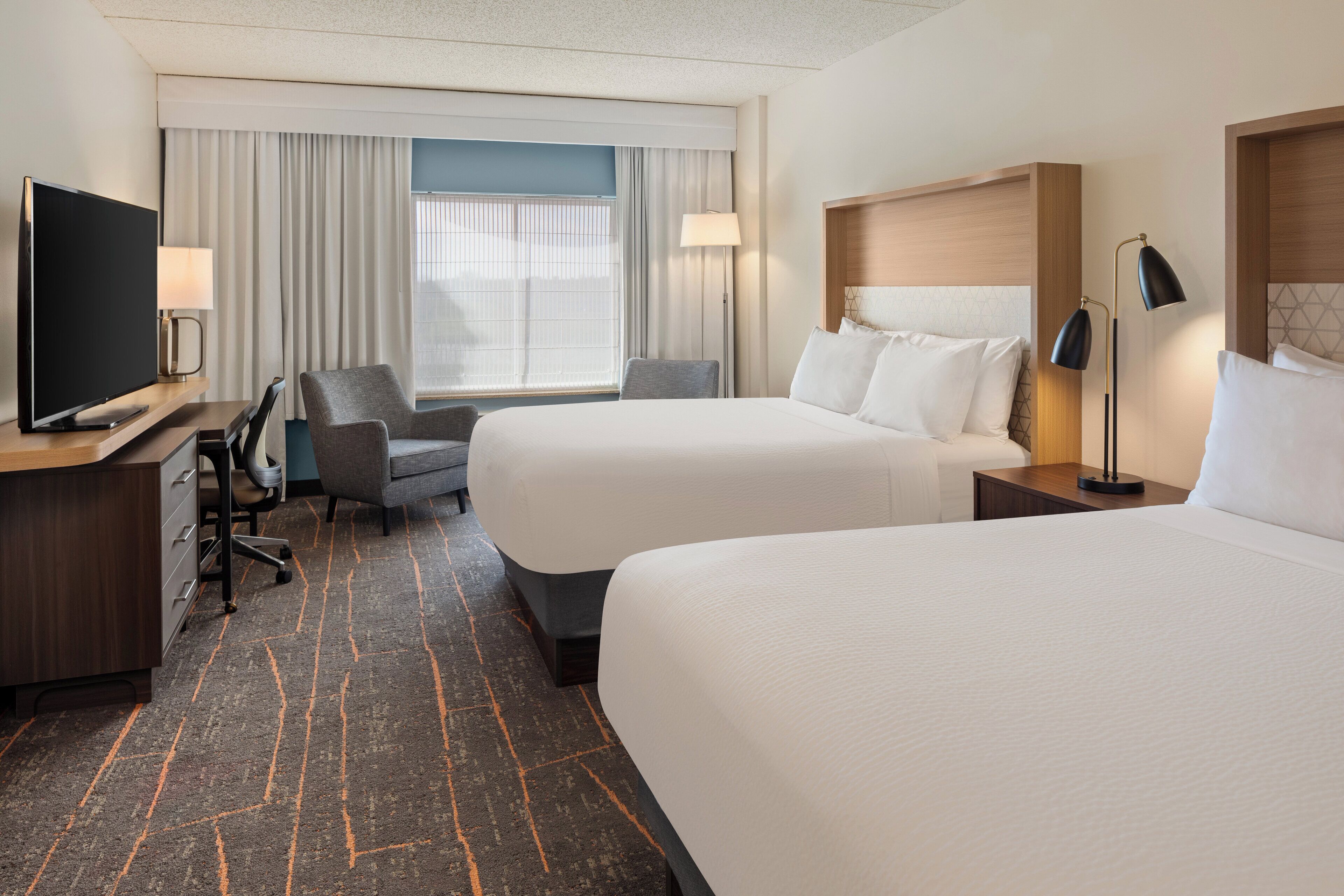 A hotel room with two beds, a desk with a TV, a chair, and a lamp.