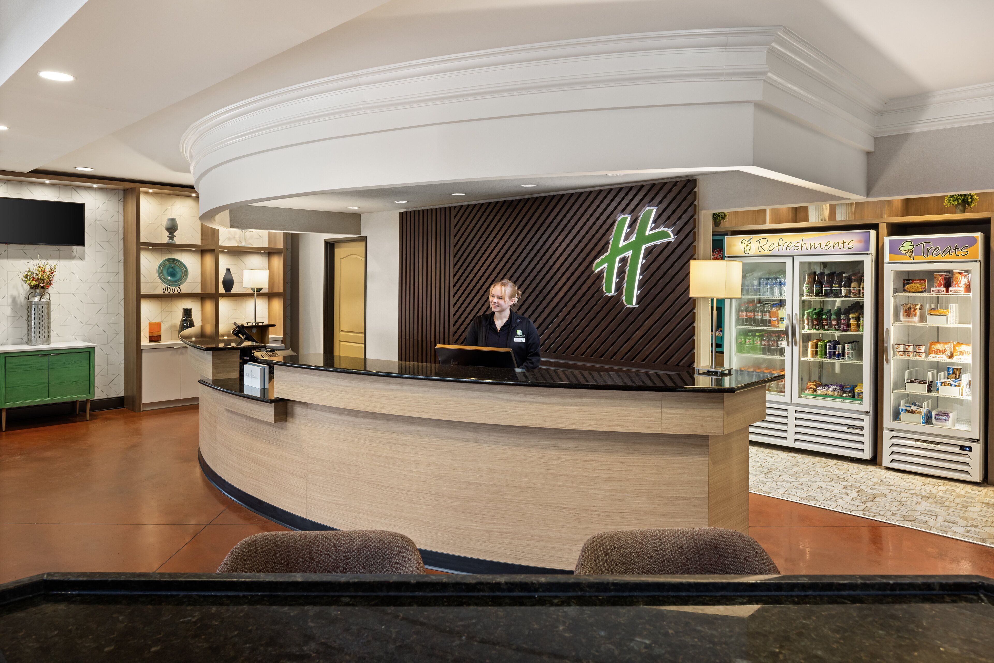 A hotel reception area with a curved reception desk, a receptionist, a wall-mounted sign, and a refrigerator with refreshments and treats.