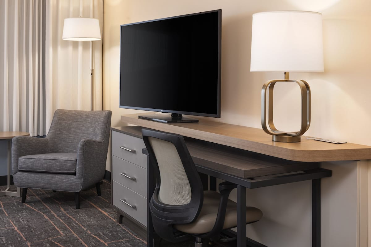 Habitación de hotel con televisión de pantalla plana, escritorio con lámpara, silla y cómoda.