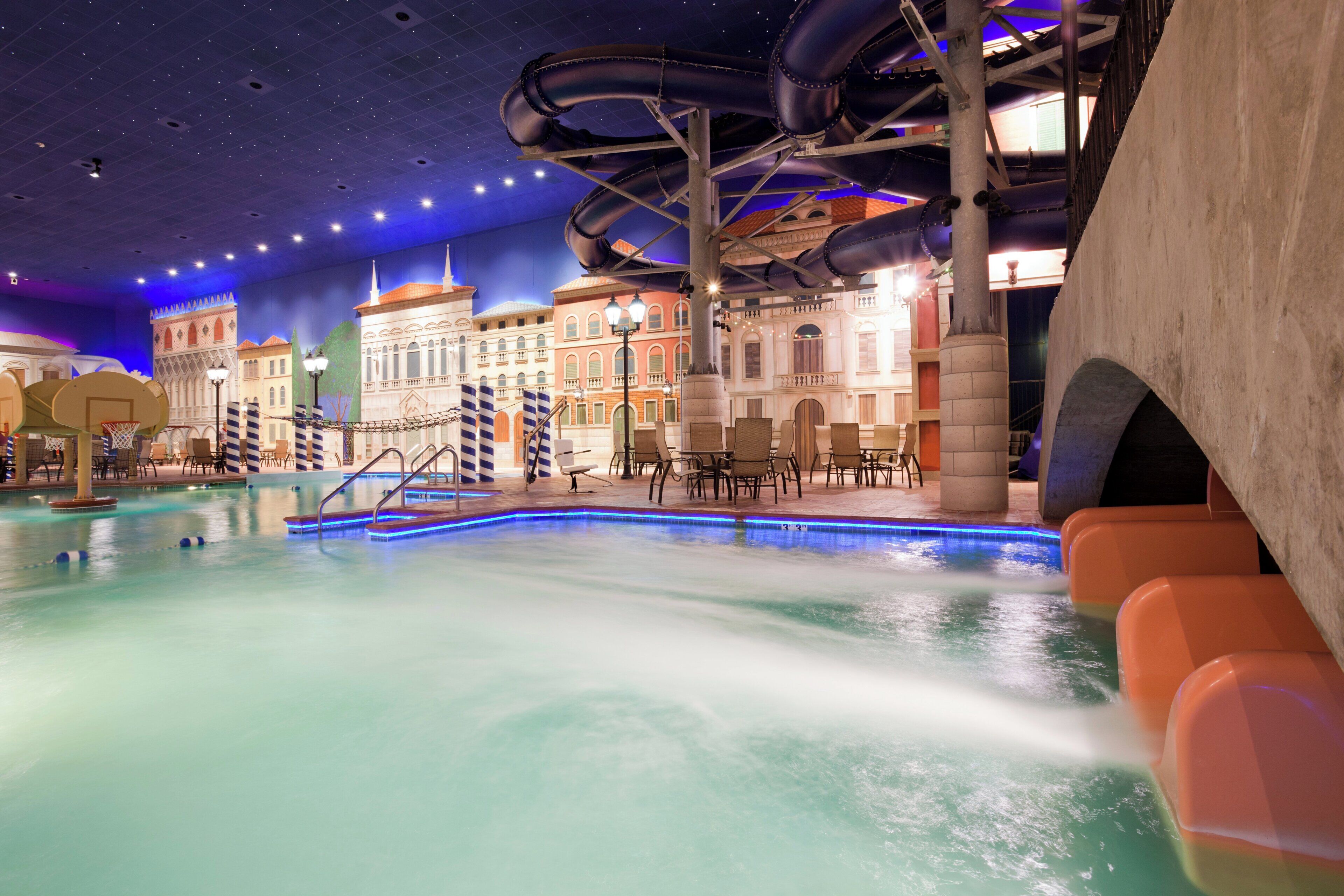 An indoor swimming pool with water slides, seating areas, and a simulated cityscape backdrop.