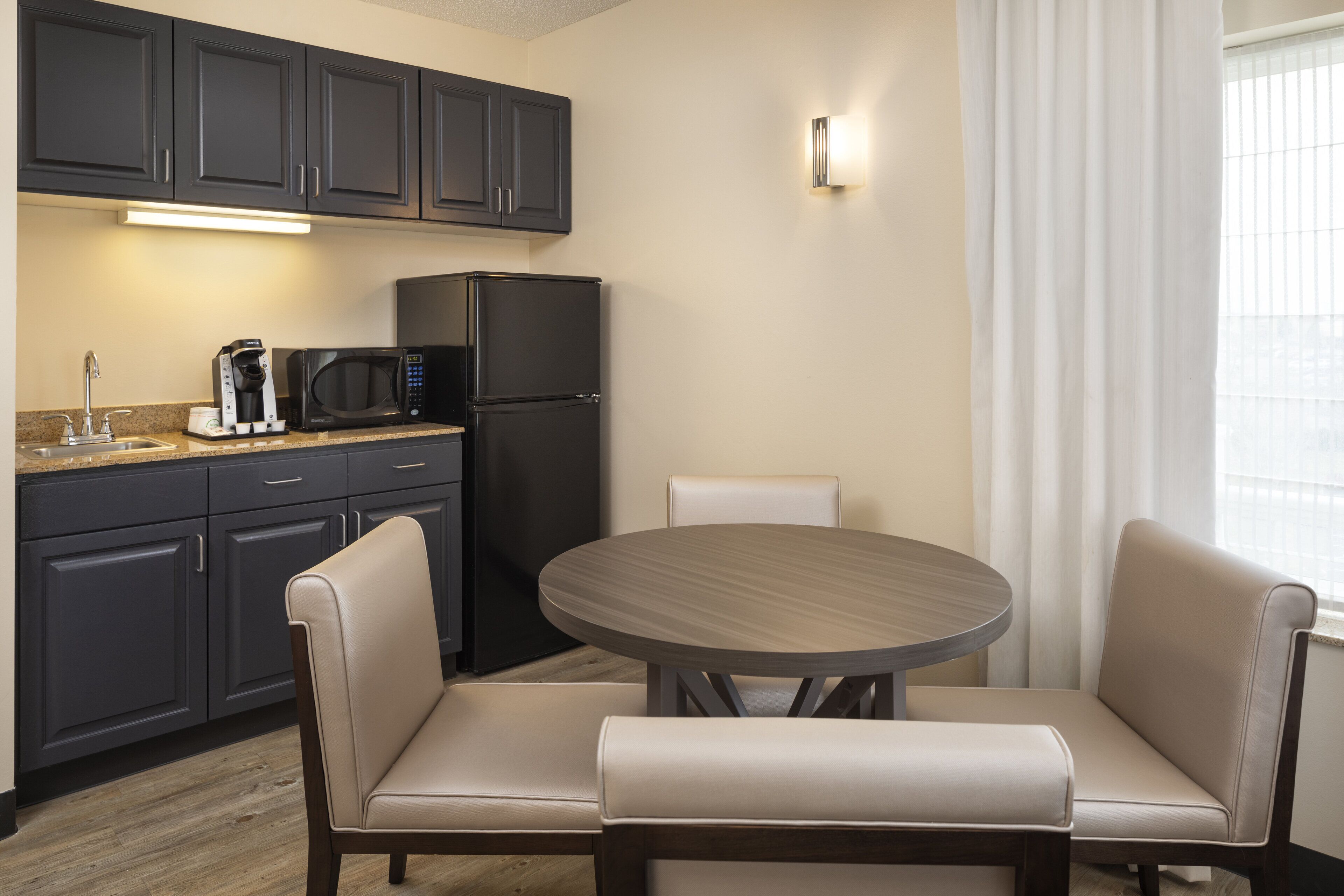 Cocina compacta con armarios oscuros, un refrigerador negro, un microondas y una cafetera. Hay una mesa redonda con dos sillas color beige.