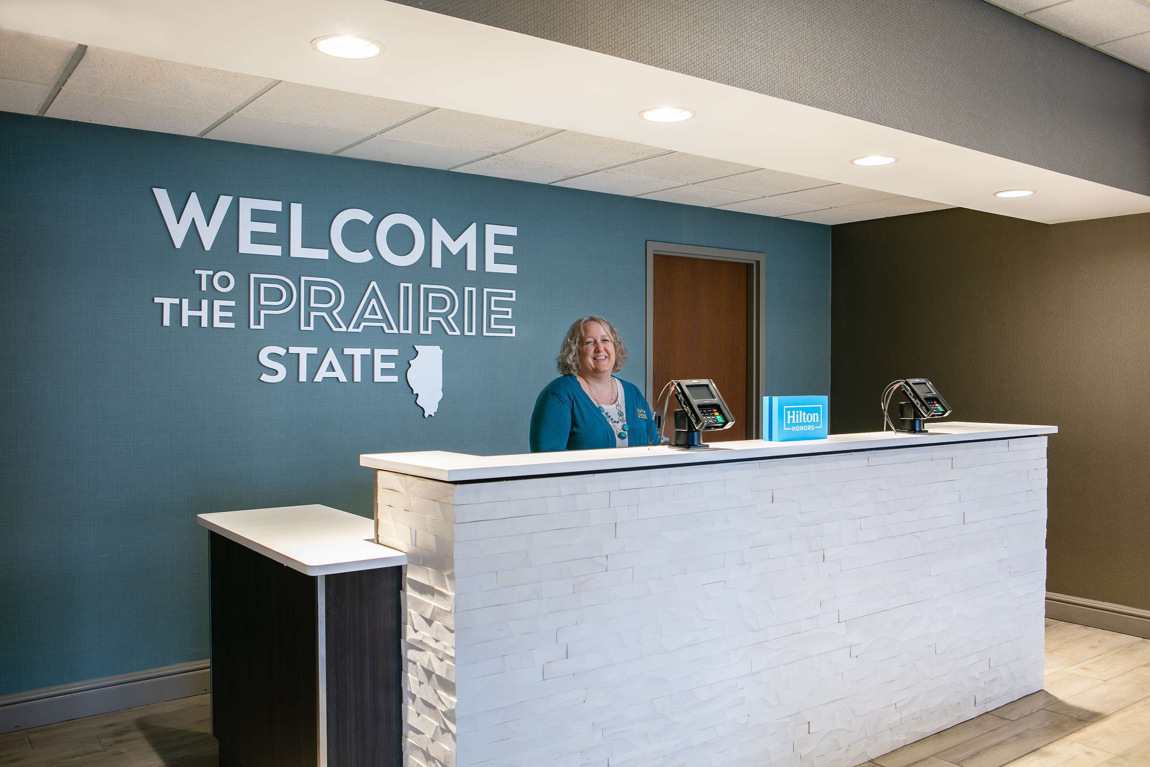 A reception desk with a "Welcome to the Prairie State" sign, a Hilton Honors card, and a digital kiosk.