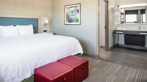 A hotel room with a large bed, a bedside table with a lamp, a small red bench, a bathroom with a mirror and sink, and a framed picture on the wall.
