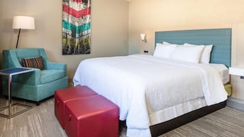 A hotel room with a bed, a chair, a red suitcase, and a painting on the wall.