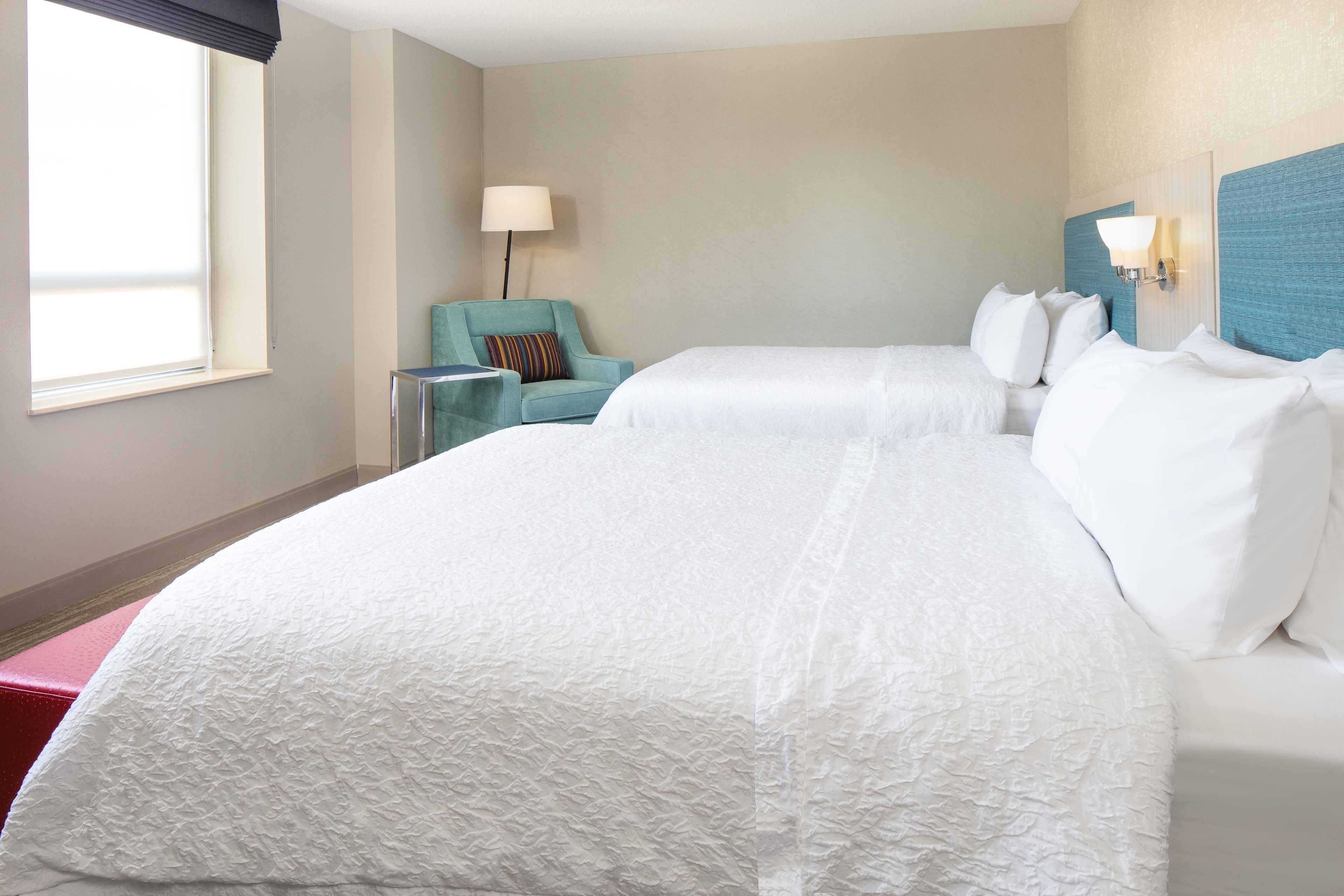 A hotel room with two beds, a chair, a lamp, and a window.