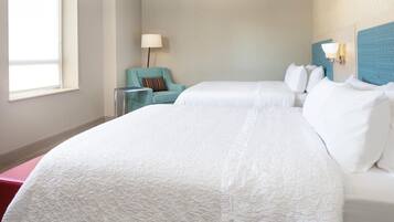 A hotel room with two beds, a chair, a lamp, and a window.