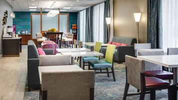 Un comedor de hotel con varias mesas y sillas, una recepción y vistas al exterior a través de amplios ventanales.