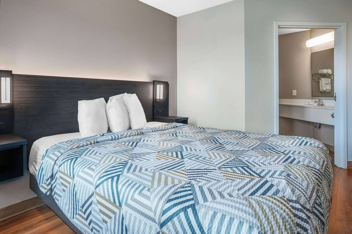A hotel room with a large bed, a nightstand, a headboard with integrated lighting, and a bathroom visible through an open door.