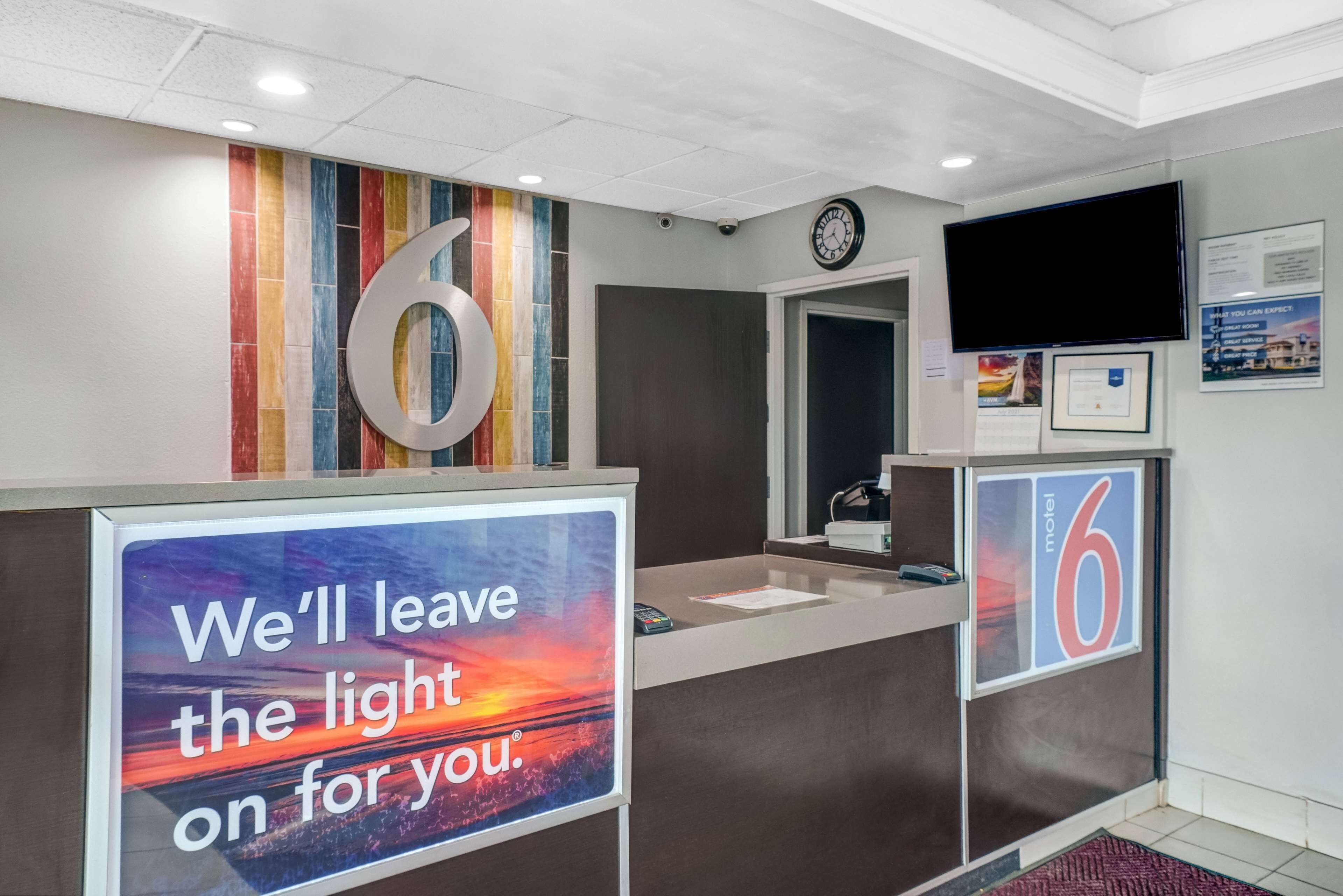 A hotel reception desk with a large number '6' sign, a clock, and a motivational sign stating "We'll leave the light on for you."