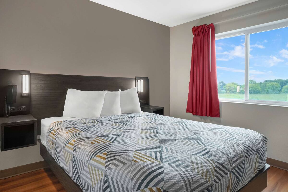 A modern hotel room with a large bed, a nightstand with a lamp, and a window with a red curtain.