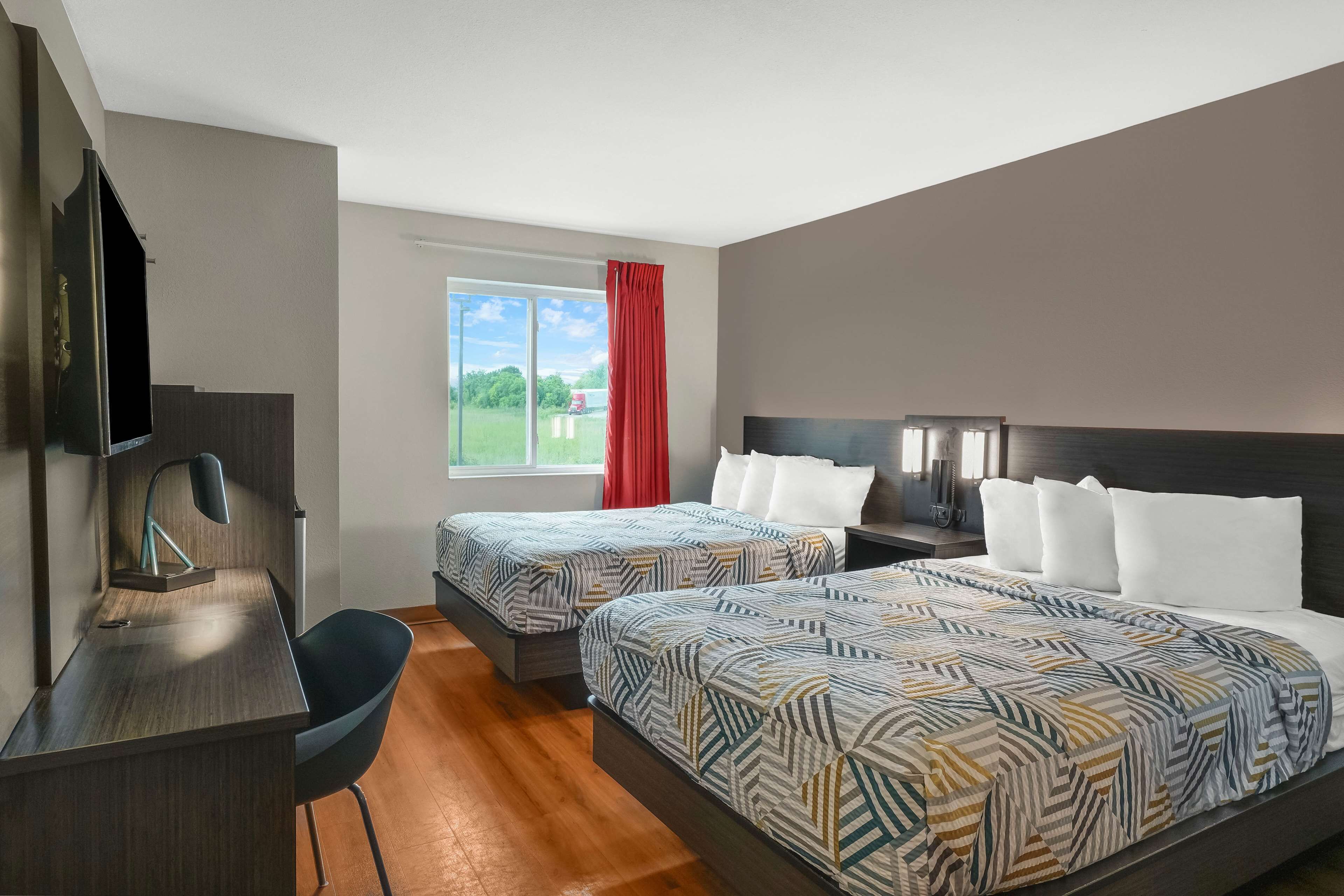 A hotel room with two beds, a desk, a chair, a TV, and a window with a red curtain.
