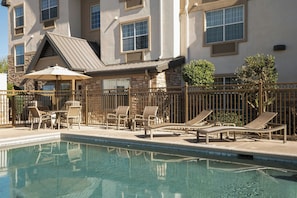 A pool area with lounge chairs and a table, surrounded by a fence, and a building with multiple windows and a sloped roof in the background.