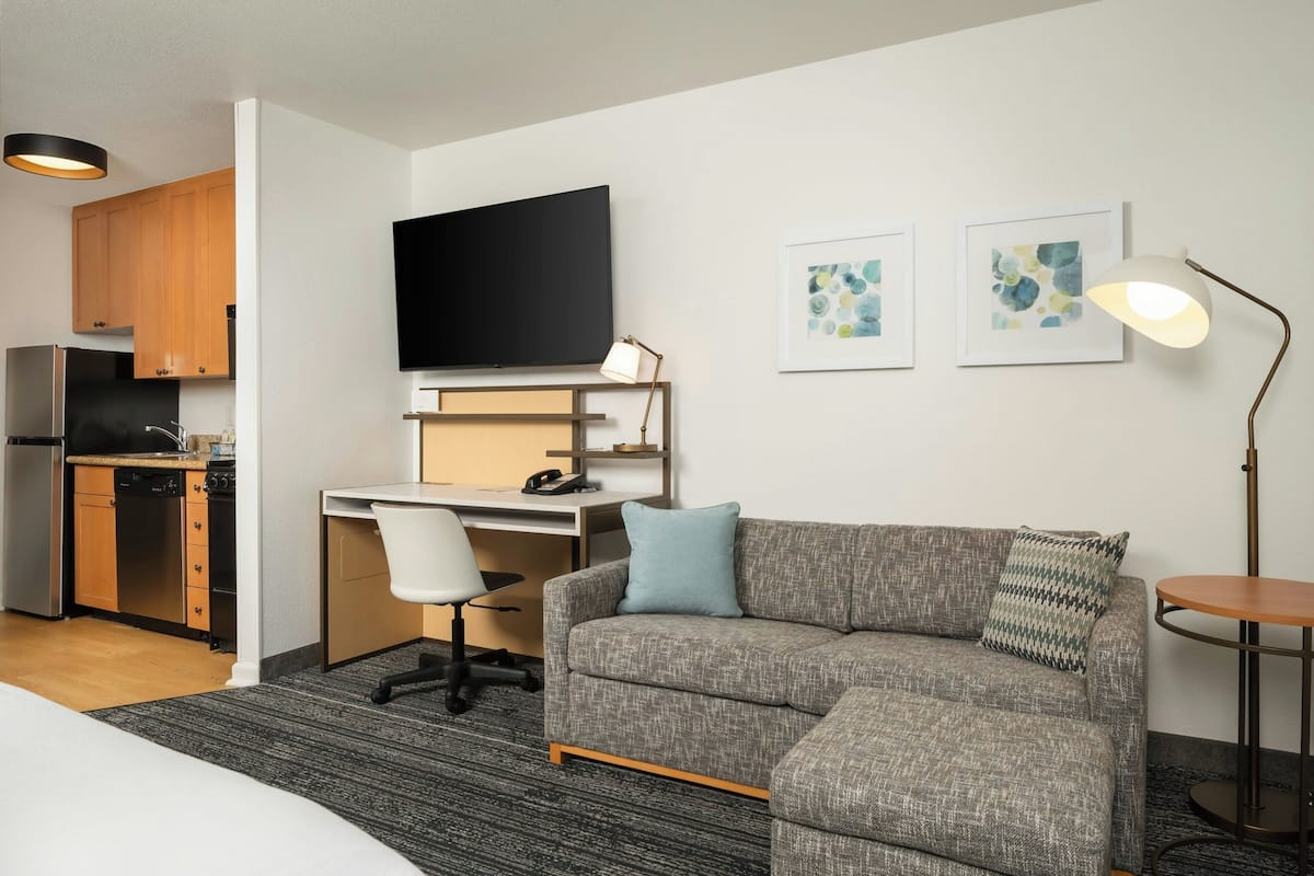 A modern hotel room with a grey sofa, a desk with a chair, a flat-screen TV, a small table, and a lamp.