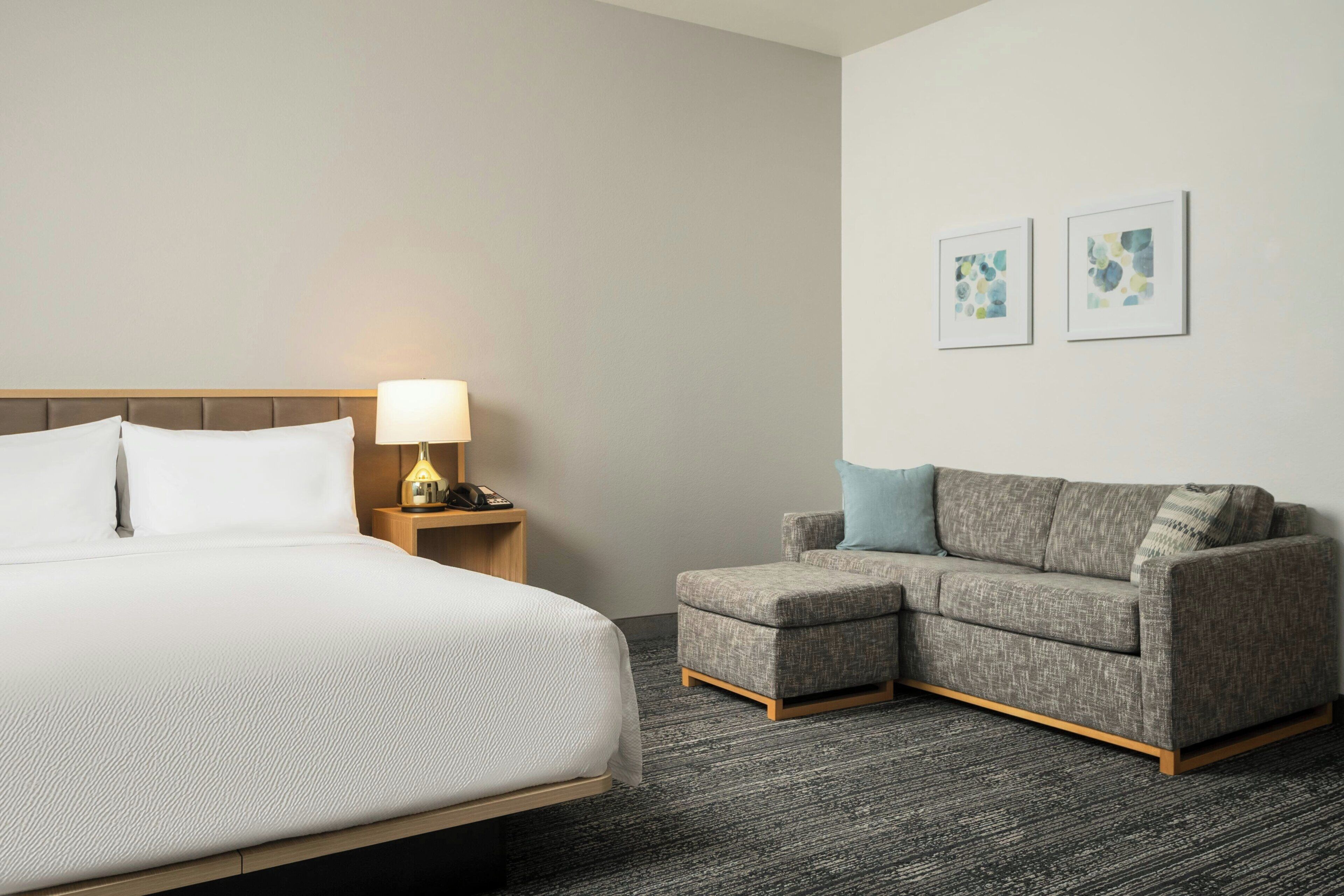 Una habitación de hotel con una cama, una mesita de noche con lámpara, un sofá gris y dos cuadros en la pared.