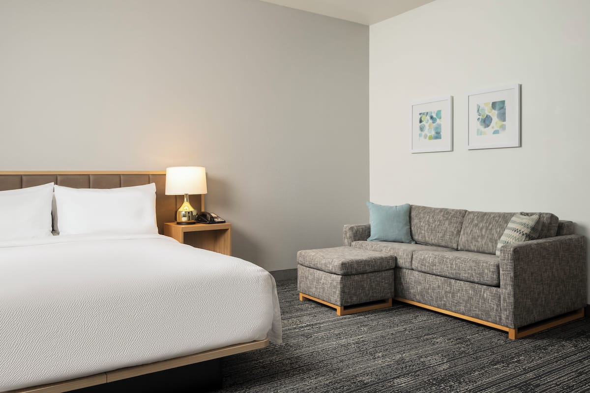 A hotel room with a bed, a nightstand with a lamp, a gray sofa, and two framed artworks on the wall.
