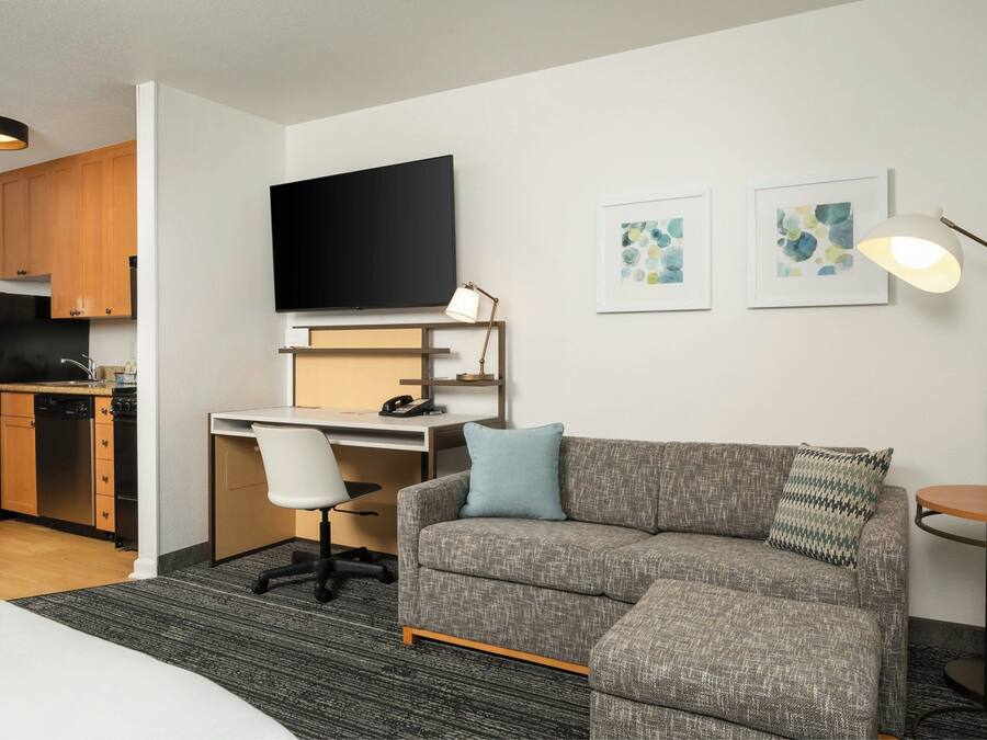 A modern hotel room with a kitchenette, a flat-screen TV, a desk, a sofa, and a lamp.