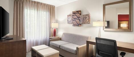 A hotel room with a flat-screen TV, a sofa, a desk with a chair, a bedside table with a lamp, and artwork on the wall.