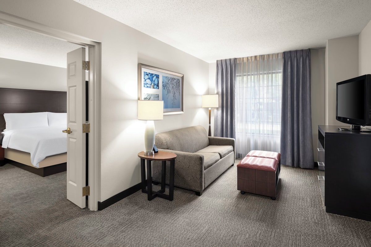 A hotel room with a bed, a sofa, a small table with a lamp, a TV, and a window with curtains.