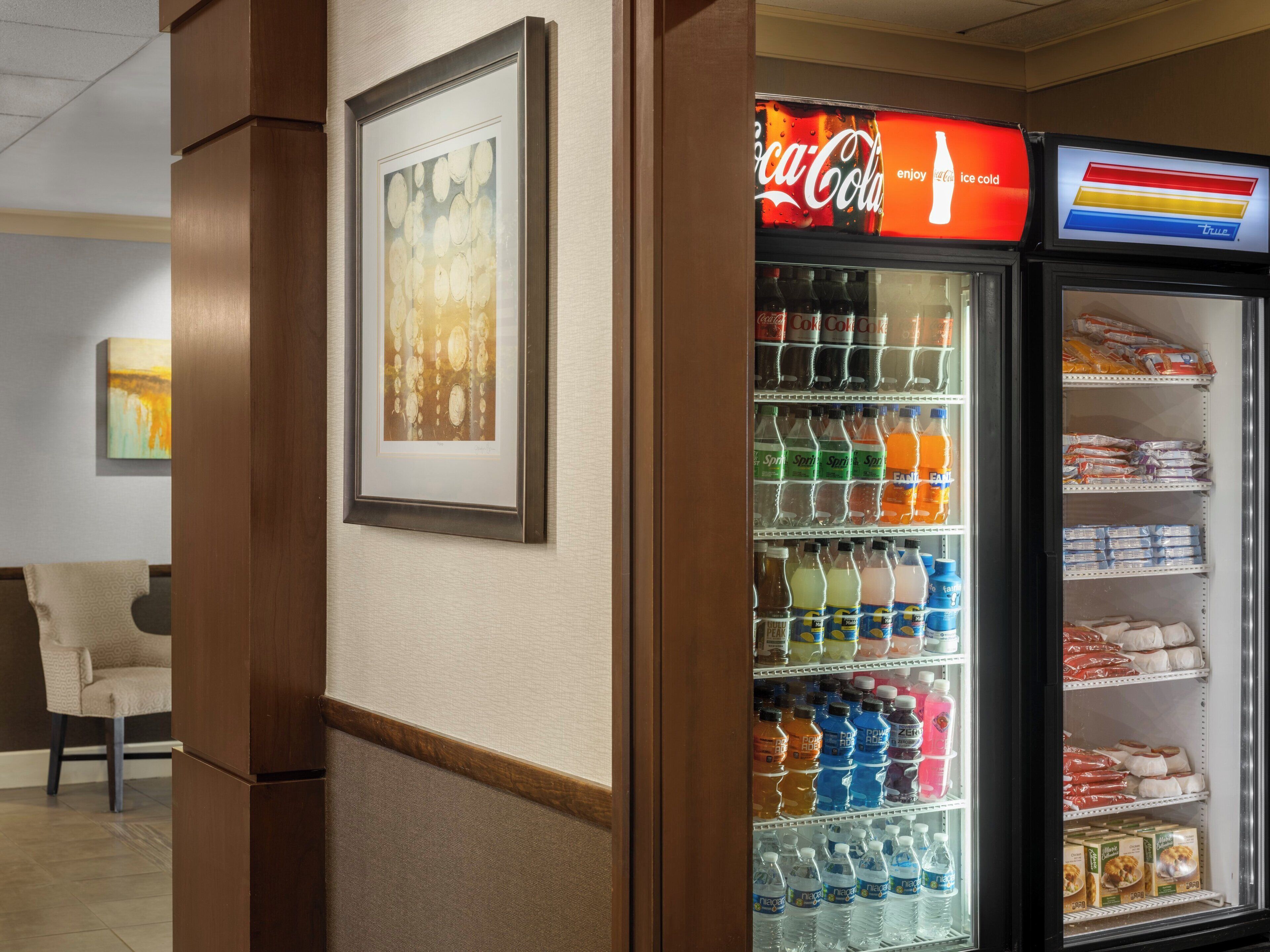 Habitación de hotel con refrigerador lleno de bebidas y aperitivos, una obra de arte enmarcada en la pared y una silla en la esquina.