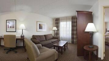 A hotel room with a brown sofa, a beige armchair, a small table, a desk with a lamp, and a large wooden cabinet.